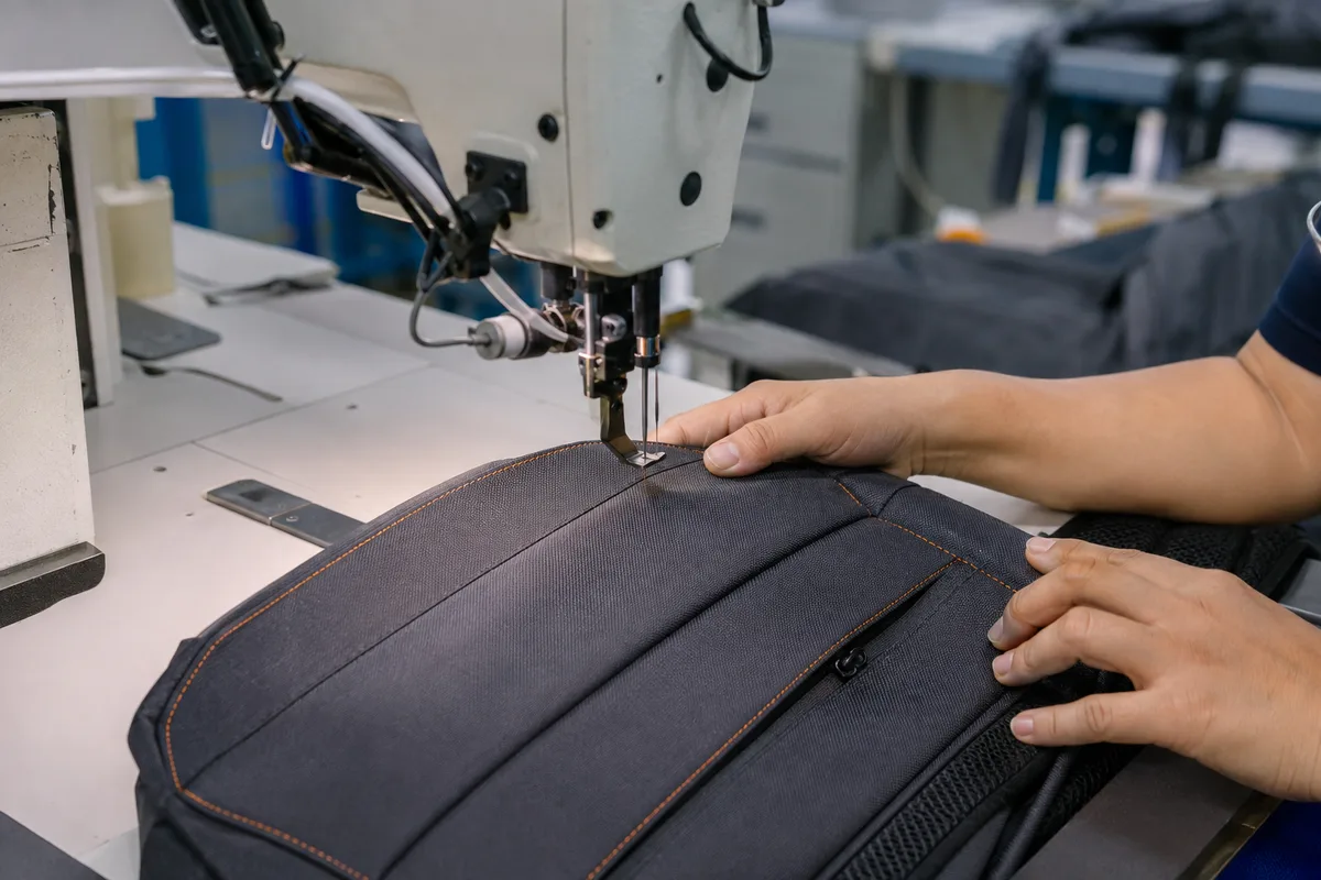 Factory worker operating an industrial Juki sewing machine stitching backpack shoulder straps, with cut fabric pieces and GRS material labels on the workbench