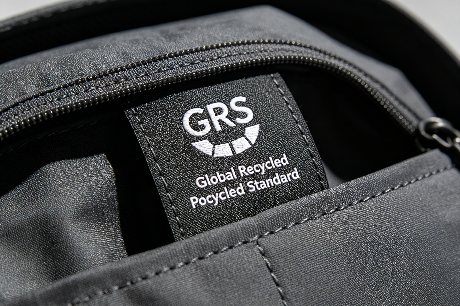 Close-up of GRS Global Recycled Standard certification woven label sewn inside a dark gray recycled polyester backpack showing label texture and stitching