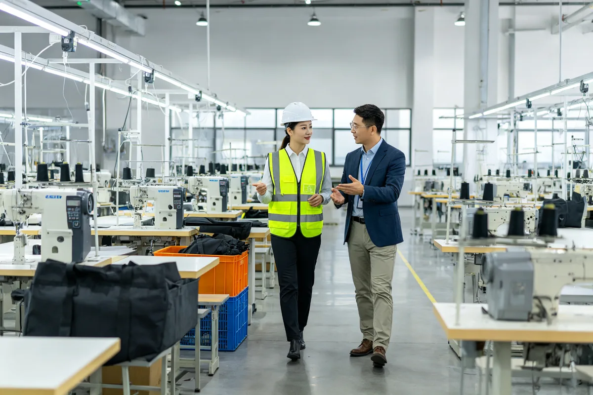 Business person in safety vest walking through a modern bag manufacturing facility with automated sewing machines and production lines during a factory tour