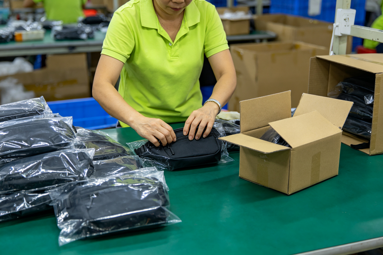 Worker placing finished black waist bags into polybags and an export carton at a small batch packaging station