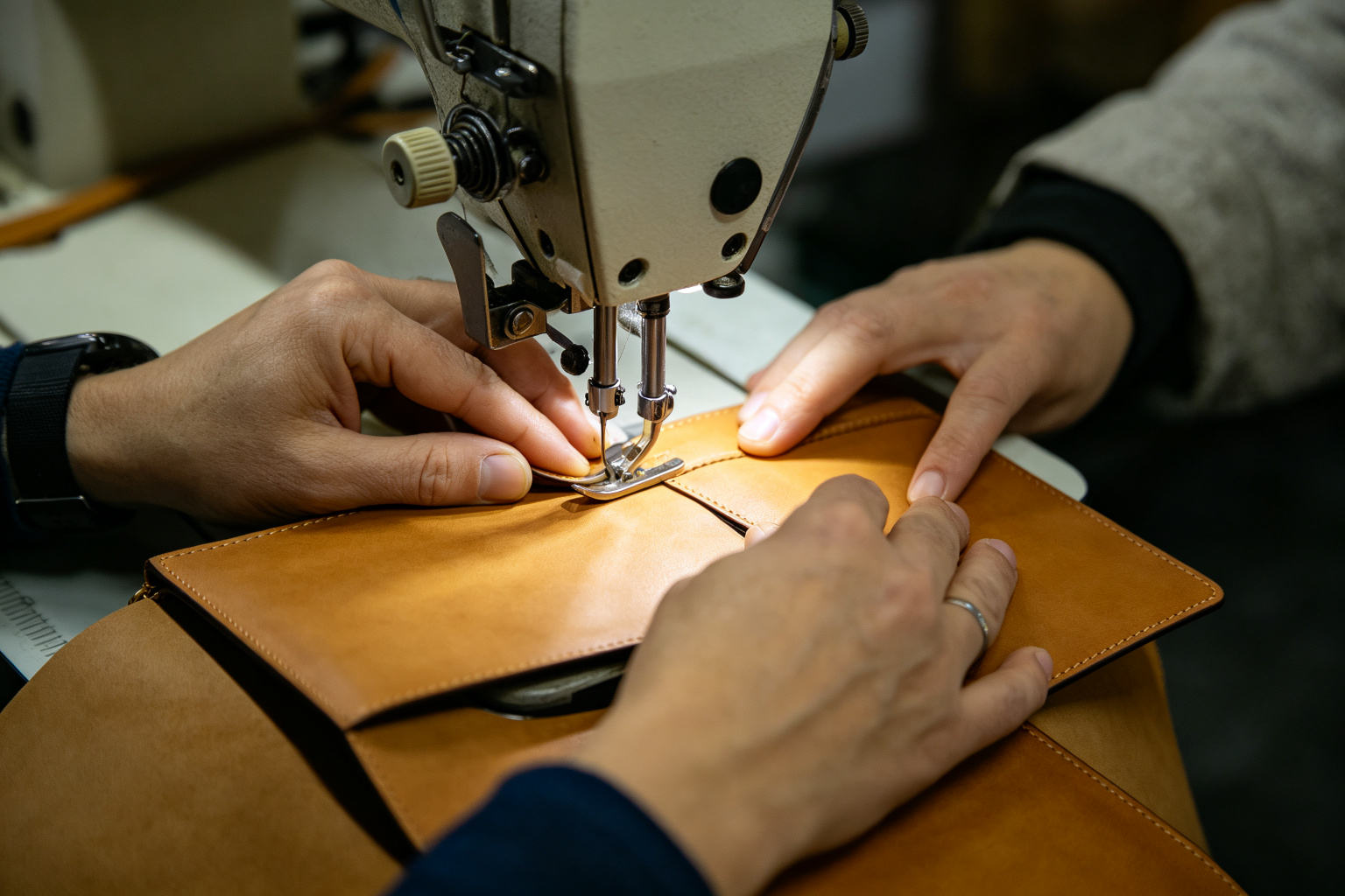 Worker sewing a tan PU leather wallet on an industrial sewing machine, hands guiding the leather under the needle