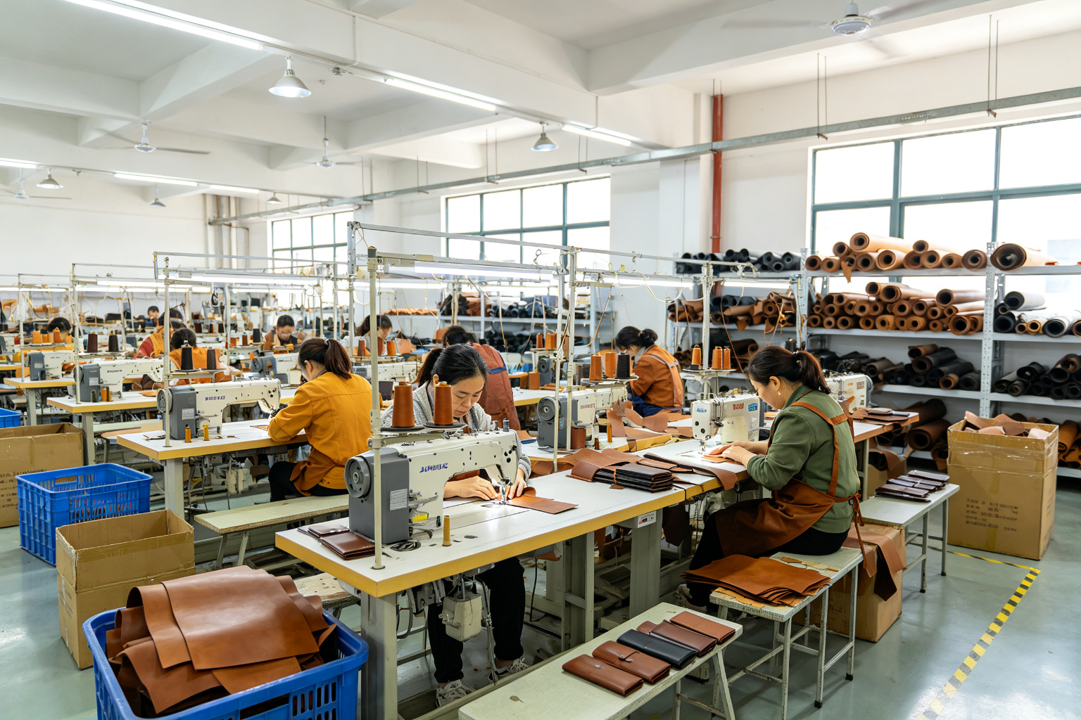 Wide angle view of a leather goods factory workshop with multiple sewing stations, leather rolls on shelves, and workers crafting PU leather wallets