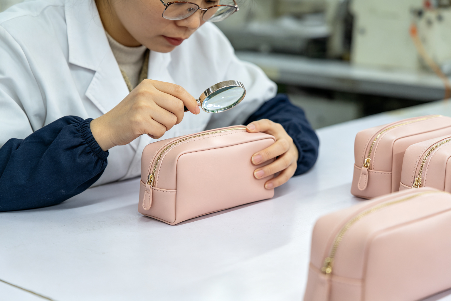 QC inspector examining a finished blush pink vegan leather cosmetic case with a magnifying glass at a quality control station