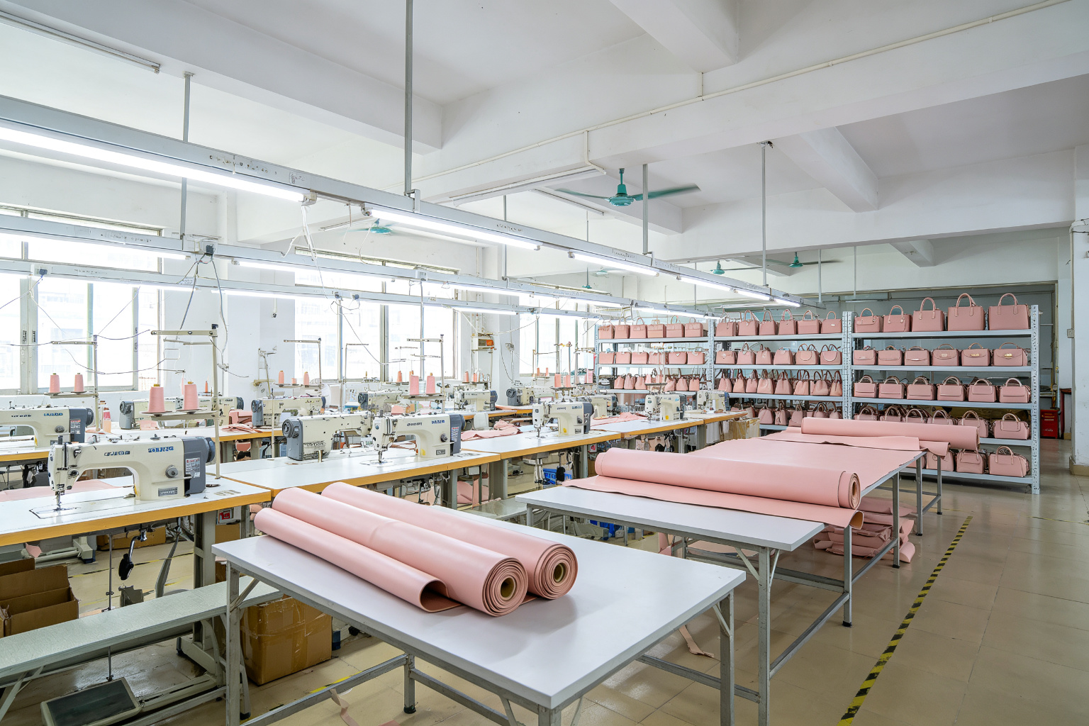 Wide angle view of a vegan leather cosmetic bag factory with sewing stations, blush pink leather rolls, and finished cosmetic cases on shelves